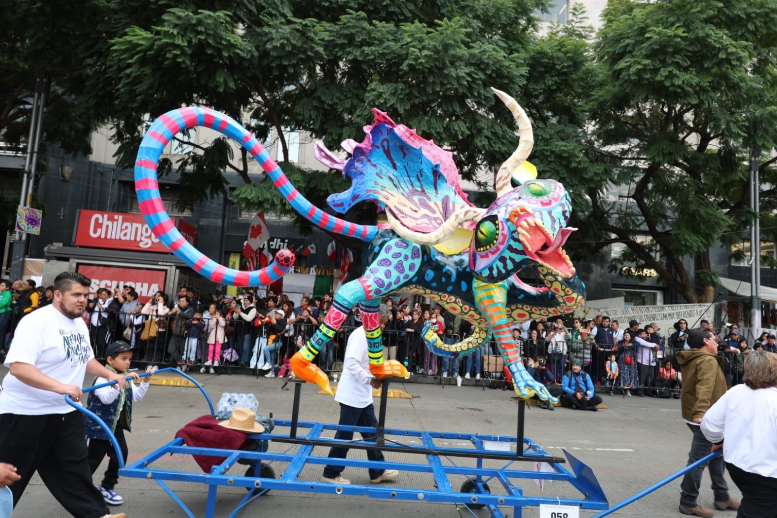 Alebrije de cartonería de grandes orejas, cola larga y vivos colores sobre carro de desfile